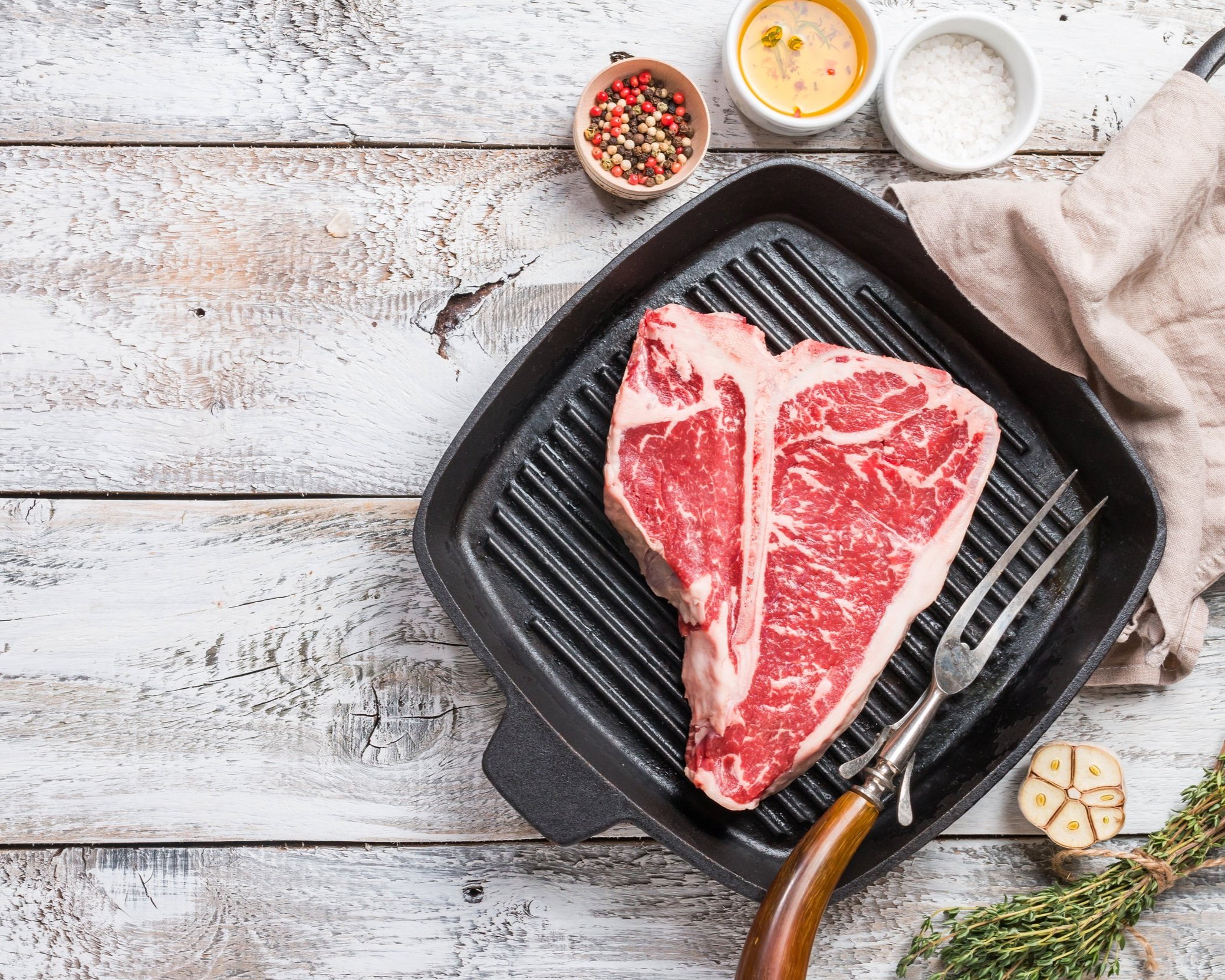 Fresh T-bone steak on a grill pan, surrounded by spices, olive oil, and garlic, ready for cooking. Ideal for gourmet meal preparation and grilling enthusiasts.