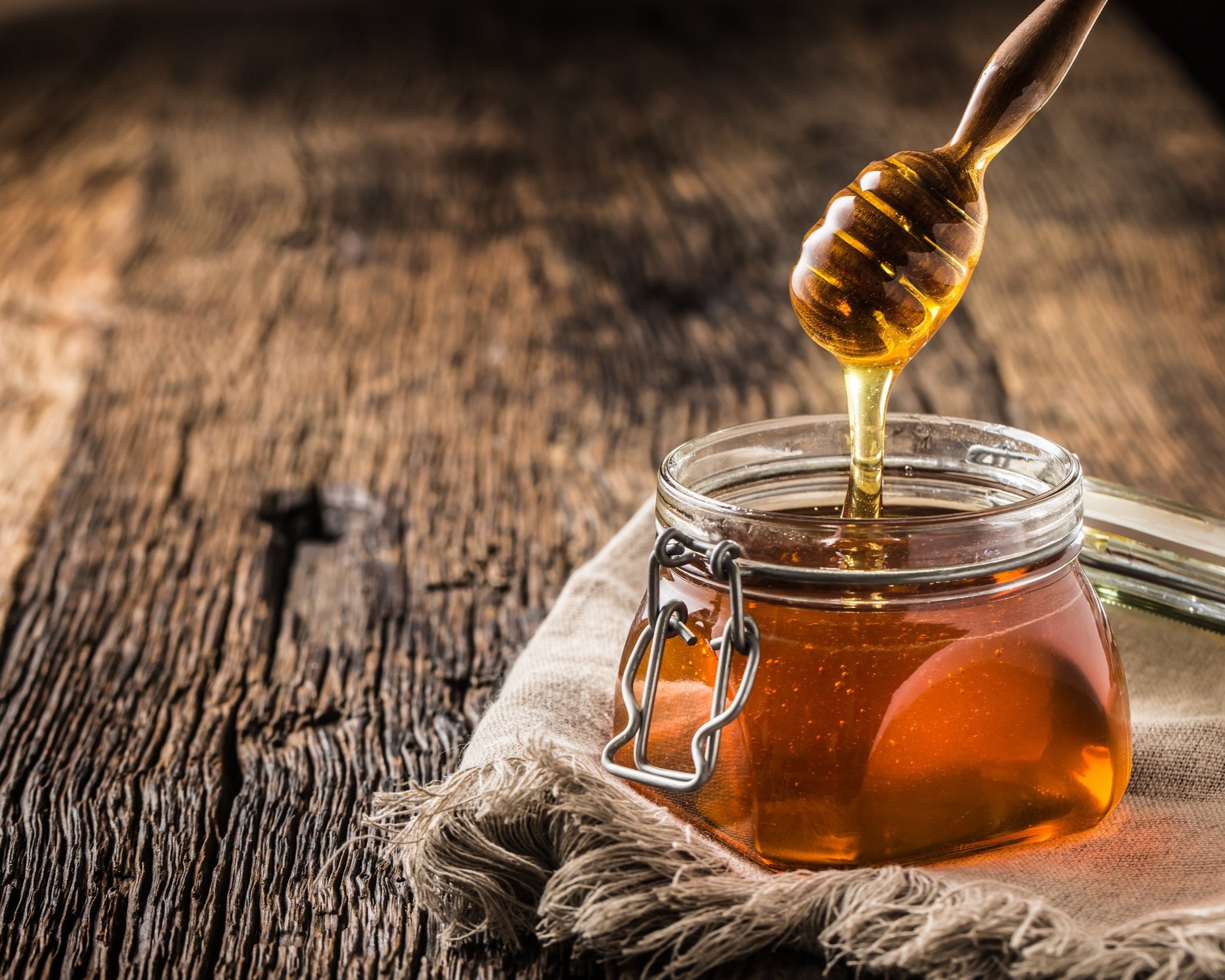 Close-up of a glass jar filled with golden honey, with a wooden honey dipper drizzling honey above it, set on a rustic wooden surface.