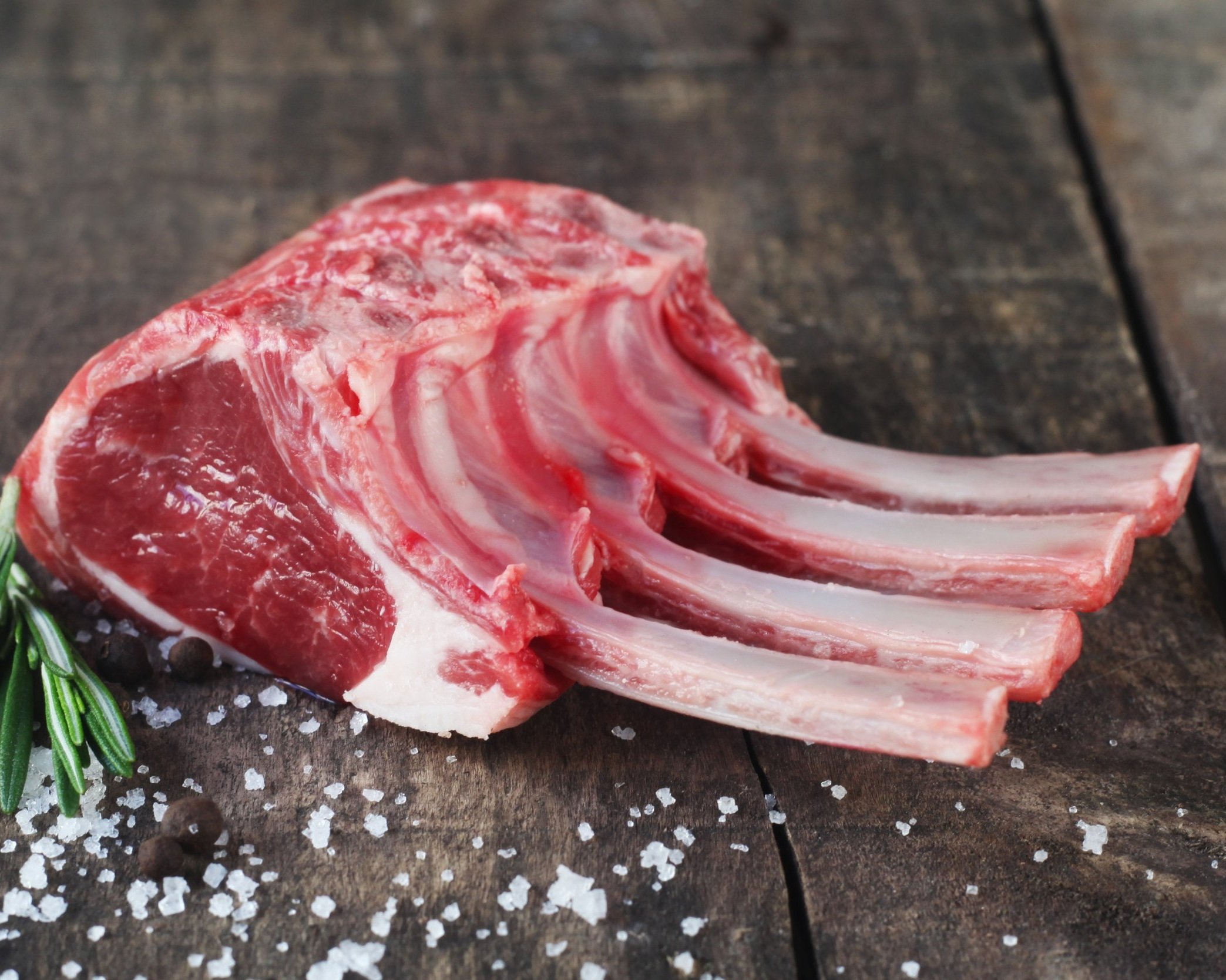Fresh lamb rack with four ribs, garnished with rosemary and surrounded by coarse sea salt and black pepper on a rustic wooden surface. Ideal for gourmet cooking and grilling.