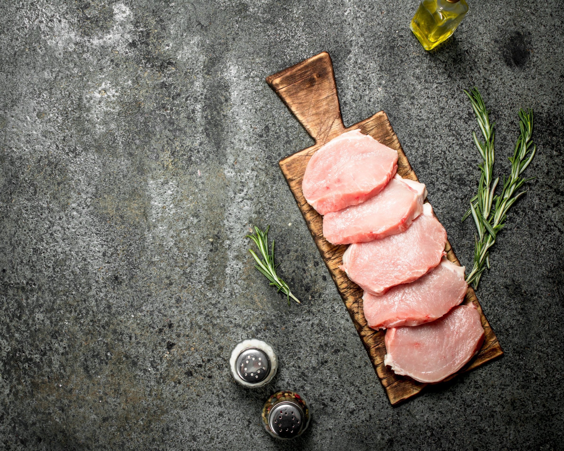 Raw pork chops arranged on a wooden cutting board, surrounded by fresh rosemary sprigs, salt and pepper shakers, and a bottle of olive oil on a textured gray surface. Ideal for cooking and meal preparation.