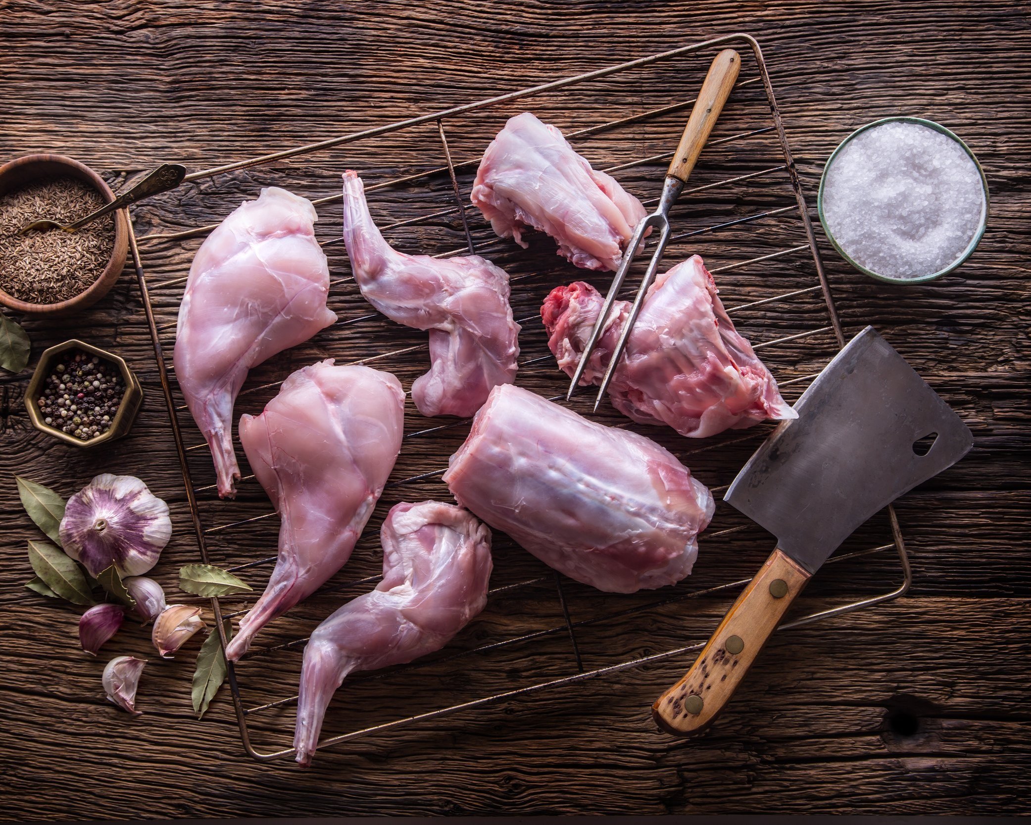 Raw rabbit meat pieces arranged on a wire rack with a butcher's cleaver, garlic, bay leaves, and spices in bowls on a rustic wooden surface.